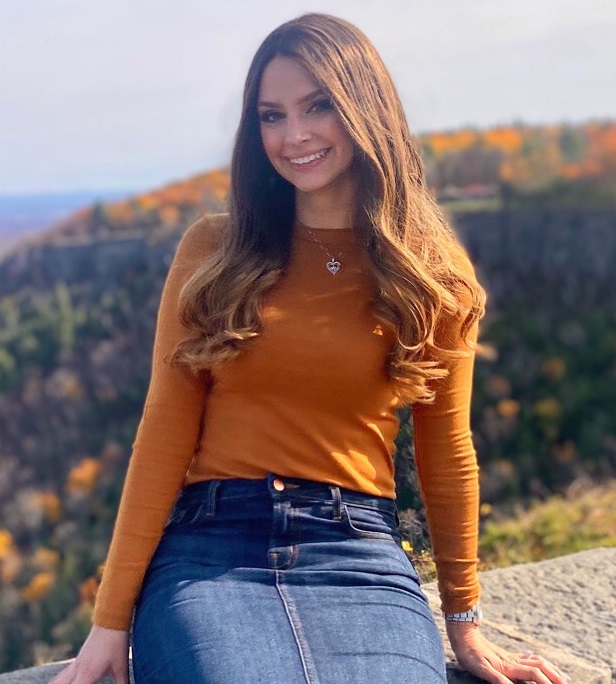 holidays: smiling woman with long brown hair, necklace, orange top, denim skirt sitting on stone wall