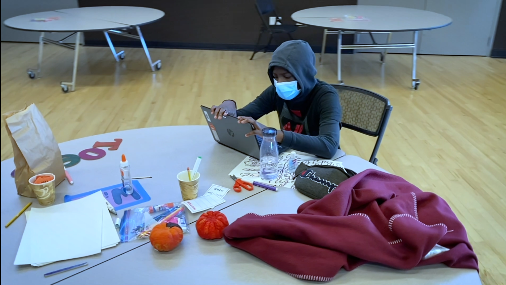  homelessness animation: Student wearing mask, hoodie sits at round table covered with laptop, school supplies 
