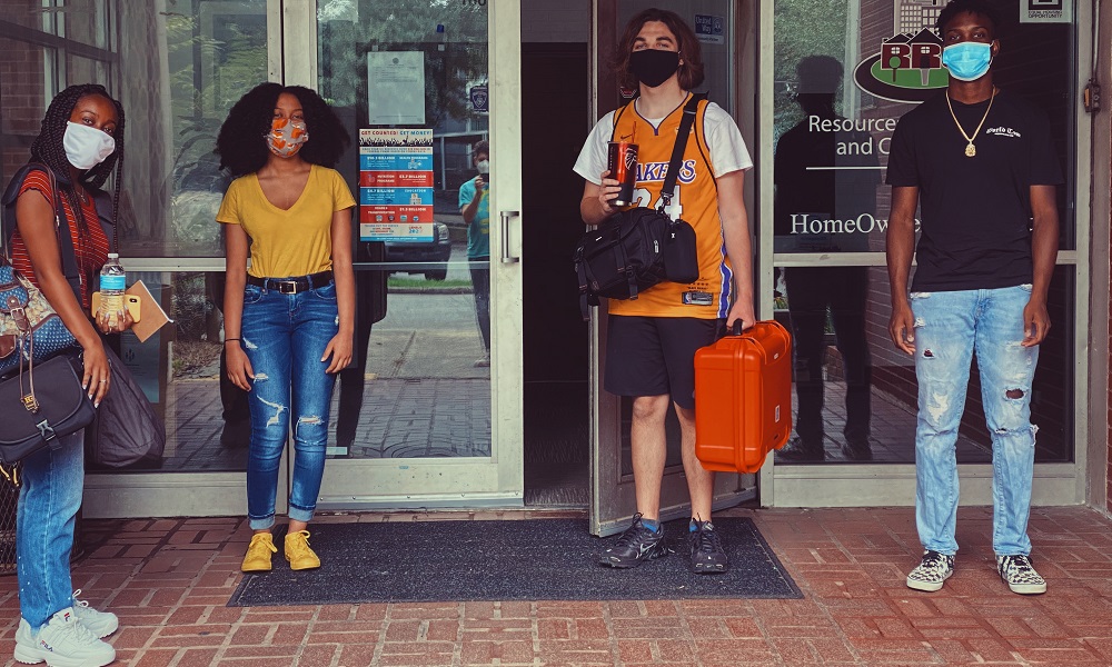 apprenticeship: 2 young women, 2 young men in face masks stand at entrance to building. One guy is carrying 2 equipment cases.