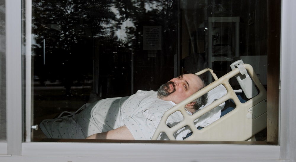 Man in hospital gown with salt and pepper hair and a goatee is seen in hospital bed through a window.