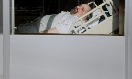 Man in hospital gown with salt and pepper hair and a goatee is seen in hospital bed through a window.