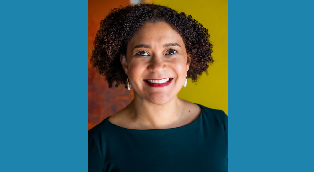 Elizabeth Lindsey newsmaker headshot; african american woman with curly hair and earings in dark blue shirt on orange background