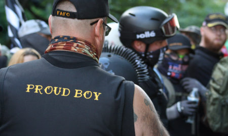 white nationalism: Rear view of man in crowd wearing ball cap, blue tank top that says Proud Boy.