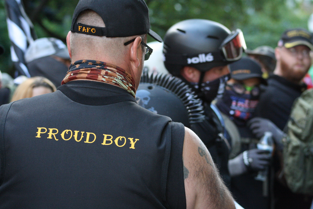 white nationalism: Rear view of man in crowd wearing ball cap, blue tank top that says Proud Boy.
