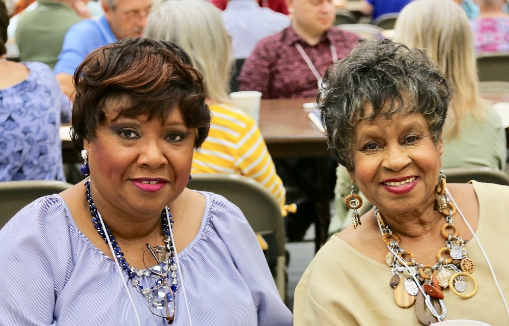 2 women, 1 smiling, with earrings, necklaces seated at long table in crowd