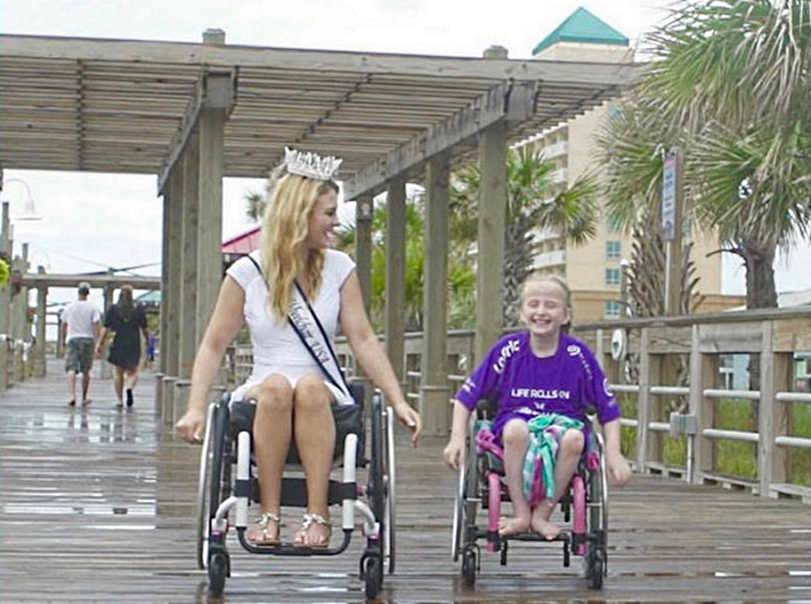 mentor: Blonde woman wearing tiara and white dress with diagonal sash in wheelchair laughs with little girl in purple T-shirt. Both are in wheelchairs on rain-soaked boardwalk.