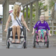 mentor: Blonde woman wearing tiara and white dress with diagonal sash in wheelchair laughs with little girl in purple T-shirt. Both are in wheelchairs on rain-soaked boardwalk.