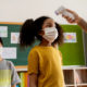 Federalfunds toschools: adult takes forehead temperature of younf girl in gold shirt with dark curly hair wearing a mask