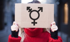 transgender athelete bill: Person in red sweater holding sign in front of face with combined gender symbol in black on cardboard