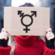 transgender athelete bill: Person in red sweater holding sign in front of face with combined gender symbol in black on cardboard