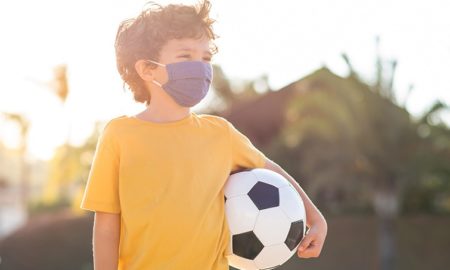 youth sports and COVID report; young child with mask on holding a soccer ball