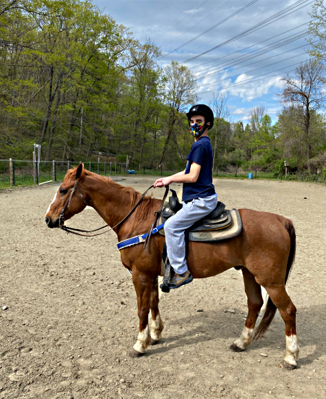 Disabled youth exercise: Clin Daly wearing blue jeans, dark t-shirt and riding helmet sits atop a chestnut horse in saddle holding reins in a dirt arean surrounded by green, leafytrees.