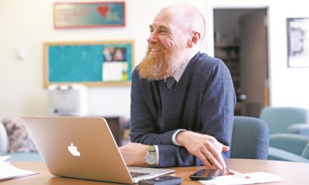 New Mexico CYFD Routinely Deletes Information That Could Be Life-Saving for Kids: Smiling man with red hair and beard in blue sweater sits with arms crossed on desk in front of laptop