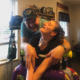 COVID vaccine access: Young woman with long blonde brais sit in wheelchair looking up at man in dark baseball cao standing behind her chair giving her a hug inside a home's living room