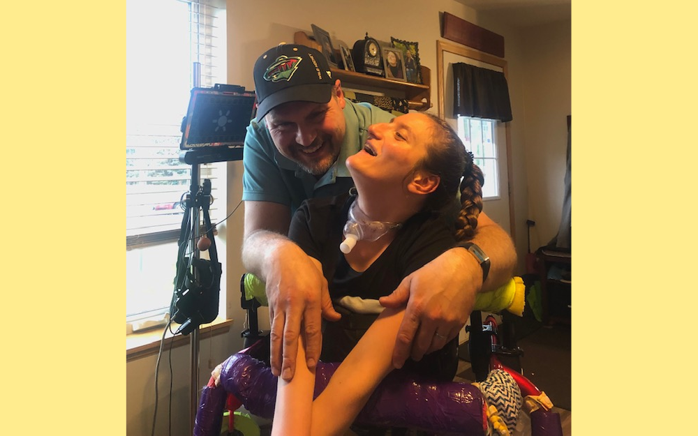 COVID vaccine access: Young woman in wheelchair with long blonde braid and colorful blanket on lap looks up at man in baseball cap standing behing her wheelchair giving her a hug