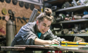 skilled trades and trades education: Yong person with brown hair in top knot leans over work tale peering at liquid in long yellow level