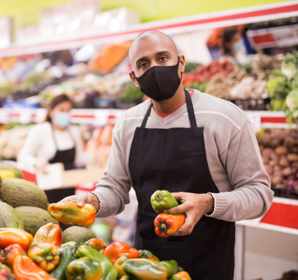 HS Internship: Young man wearing full, dark apron and face mask stacking produce in a grocery display