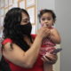 Payments cut child poverty: Dark- haired woman wearing black mask and red sleeveless top stands holds dark-haired baby wearing red outfit on her left hip.