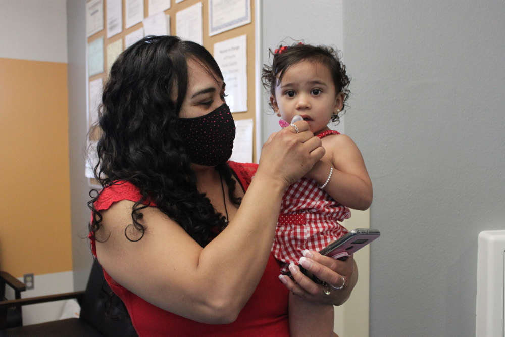 Dark- haired woman wearing black mask and red sleeveless top stands holds dark-haired baby wearing red outfit on her left hip.