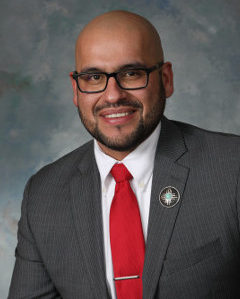 Child poverty: Rep. Javier Martínez headshot - smiling bald man with dark beard wearing dark-framed glasses, gray suit, white shirt & red tie against grey background