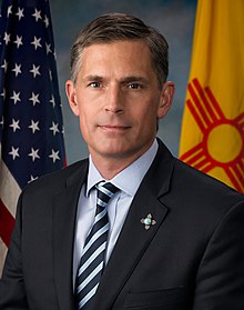 Child poverty: Sen. Marin Heinrich headshot dark-haired man wearing navy suit, light blue shirt and navy with silver stripped tie in front of American and New Mexico flags 