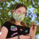 Young girl in black t-shirt wearing a mask, holds up small brown elephant sculpture in left hand, standing in front of leafy green tree