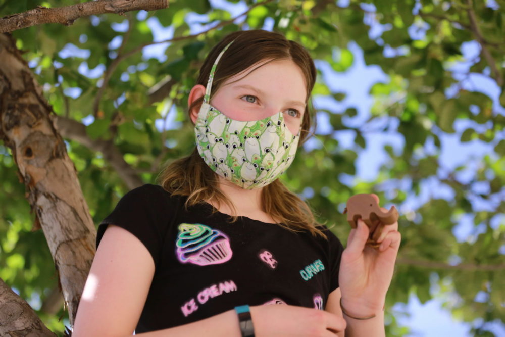 Young girl in black t-shirt wearing a mask, holds up small brown elephant sculpture in left hand, standing in front of leafy green tree
