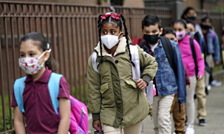 Pandemic academic damage: Four elementary students wearing maks walk spaced out, single file along a brown wood fence.
