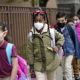 Pandemic academic damage: Four elementary students wearing maks walk spaced out, single file along a brown wood fence.