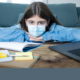 NY mental health school budget cuts: Ypung, dark-haired girl in light blue shirt wearing mask, sits at desk with head on arms