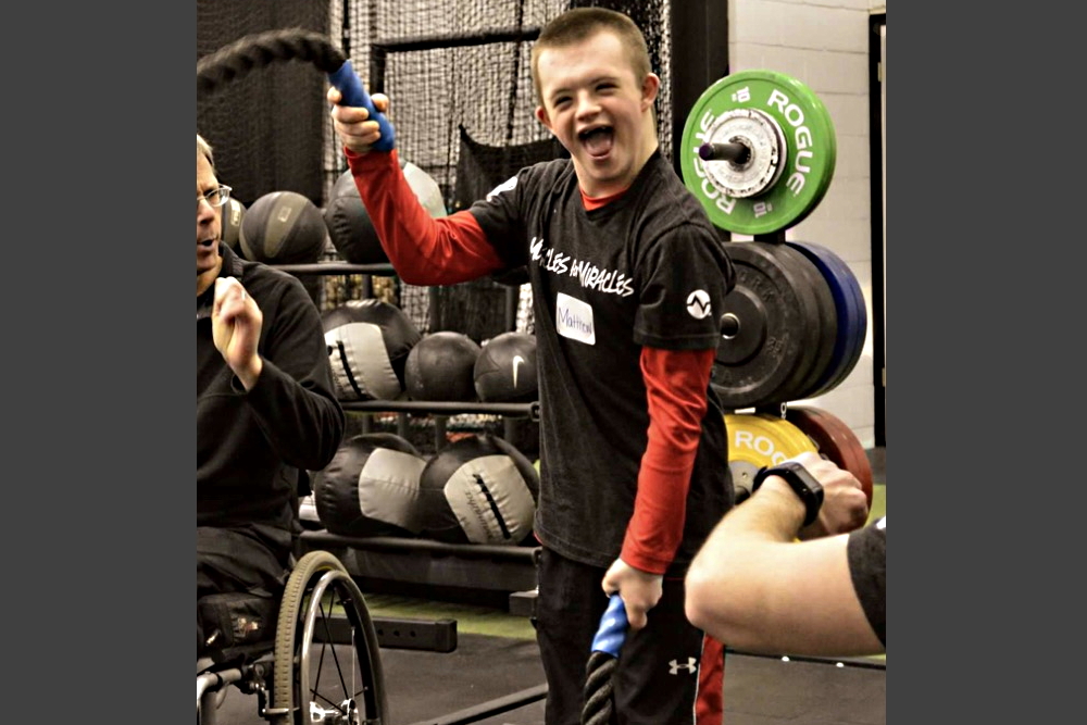 Disabled youth fitness: young man in black and red gym clothes holds workout rope standing in frong of a free weights set and next to another person in a wheelchair in a gym.
