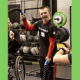 Disabled youth fitness: young man in black and red gym clothes holds workout rope standing in frong of a free weights set and next to another person in a wheelchair in a gym.