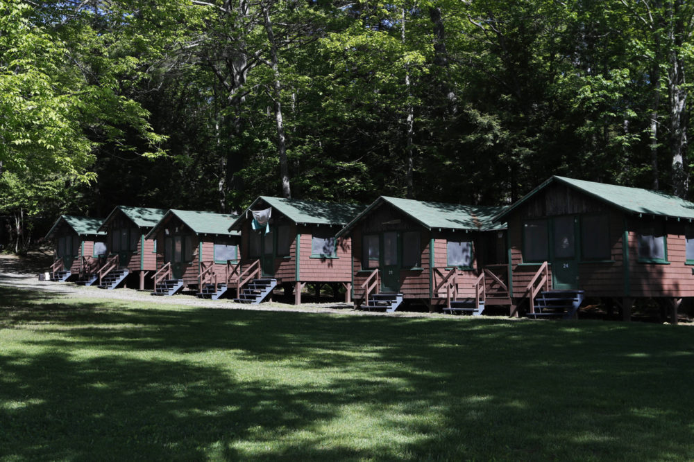 CDC Camp Mask mandates: Row of rustic brown cabins with green roofs,on lawn surrounded by dense, tall trees