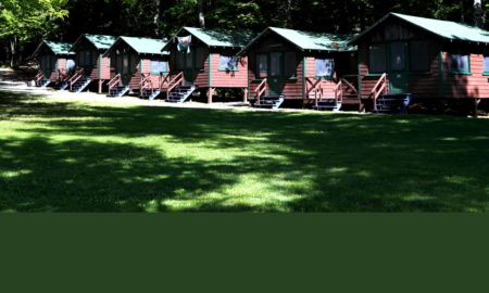 CDC Camp Mask mandates: Row of rustic brown cabins with green roofs,on lawn surrounded by dense, tall trees
