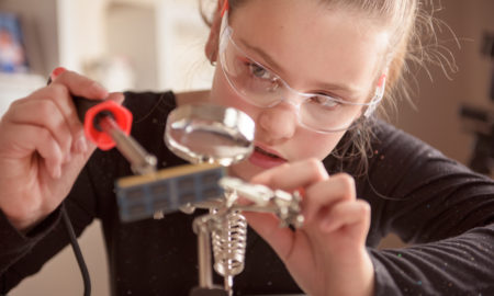 Virtual summer camps: Little girl with safety glasses works with a soldering iron on a computer component