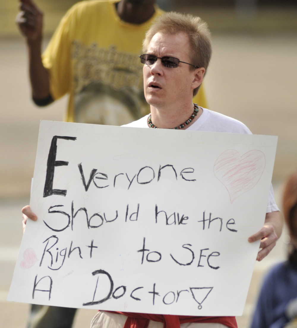 Alabama Medicaid expansion: Ray ables, man with blond hair and sunglasses holding wite sign with black marker text "Everyone should have the right to see a doctor."