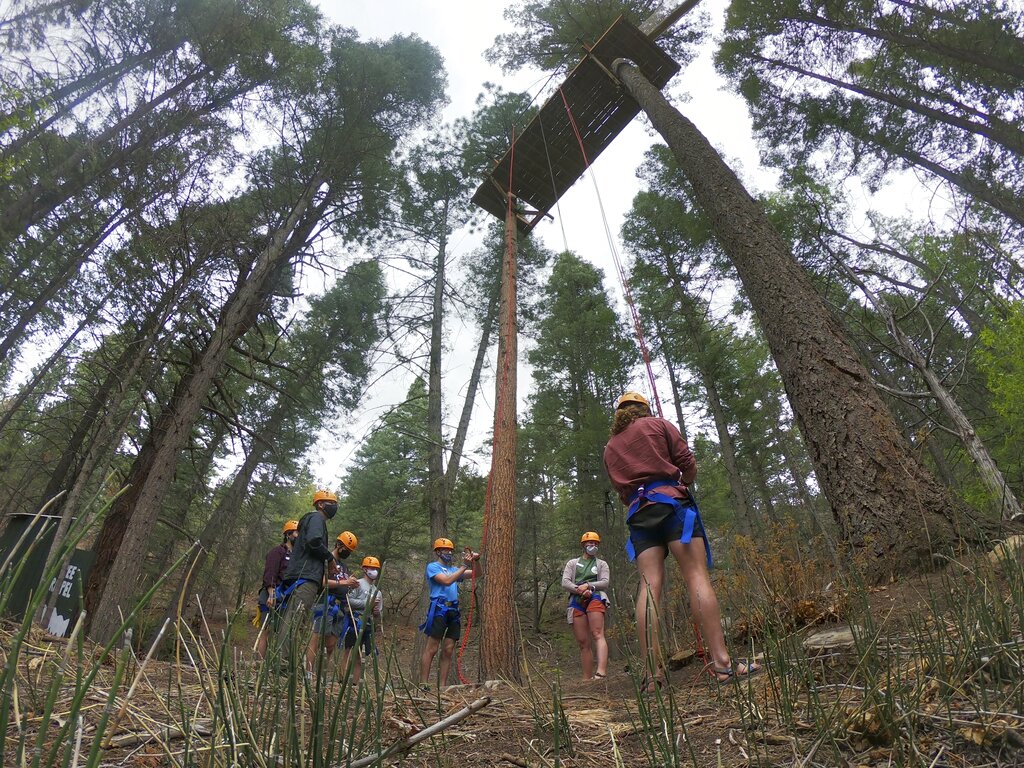 COVID Camp guidelines: Campers wearing shorts, t-shirts, facemasks, yellow hardhats and rappelling gear stand amidst tall trees beneath a rapelling platform high in the trees.