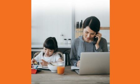 2021 KIDS COUNT Data Book: Asian mother working at kitchen table on laptop with daughter doing homework and eating beside her