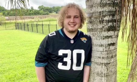 Mental health Kids Isolation Reentry: Young man with blonde, curly hair wearing black #30 football jersey leans against palm tree trunk in green grassy field