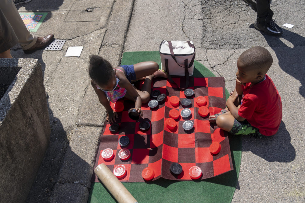 Playstreets Philadelpha: Two young children sit on the street playing chckers on an over-sized red and black checkered game board.