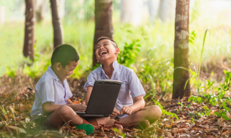 Summer SEL: Two young Asian boys in light blue shirtssit on forest floor with a laptop laughing.