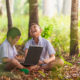 Summer SEL: Two young Asian boys in light blue shirtssit on forest floor with a laptop laughing.