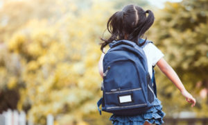 American rescue plan funds flow to summer OST programs: small girl with dark hairin pigtails and blue backpack wearing ruffled blue skirt and white top walks away from camera