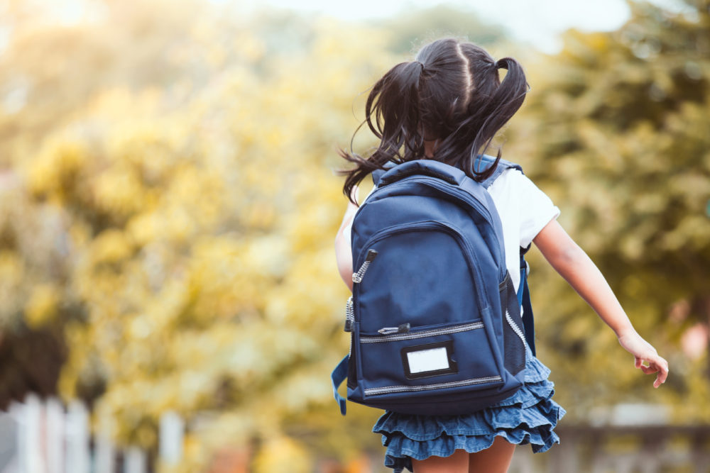 American rescue plan funds flow to summer OST programs: small girl with dark hairin pigtails and blue backpack wearing ruffled blue skirt and white top walks away from camera