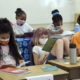 School funding addresses summer program disparities: Six masked elementary-age children sit at two rows of desks reading and writing