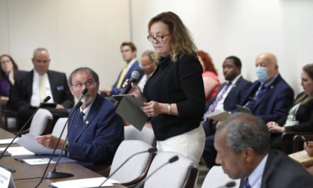 Child Marriage-North Carolina: Woman stands speaking at roundtable with microphones surrounded by about a dozen seated people in white room
