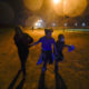 Migrant children: 3 masked children run in the rain silhouetted against a street-lamp lit dark sky