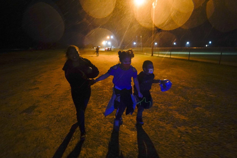 Migrant children: 3 masked children run in the rain silhouetted against a street-lamp lit dark sky