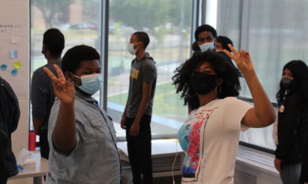 Afterschool programs use data to meaure outcomes: Eight masked teens stand in white room with large windows; two black girls in front flashing peace sign with hands