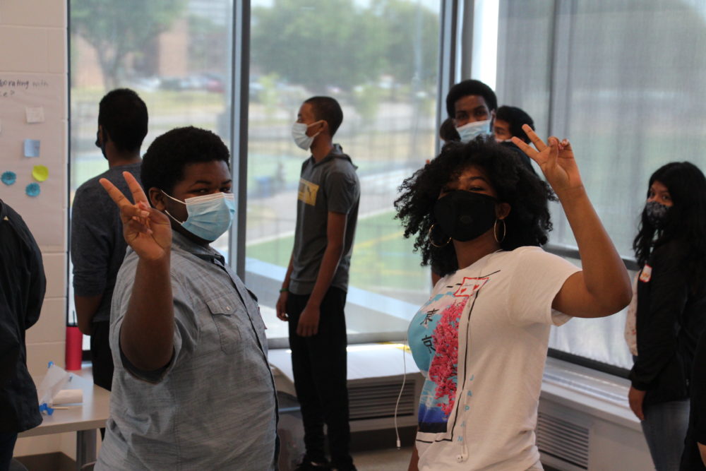 Afterschool programs use data to meaure outcomes: Eight masked teens stand in white room with large windows; two black girls in front flashing peace sign with hands